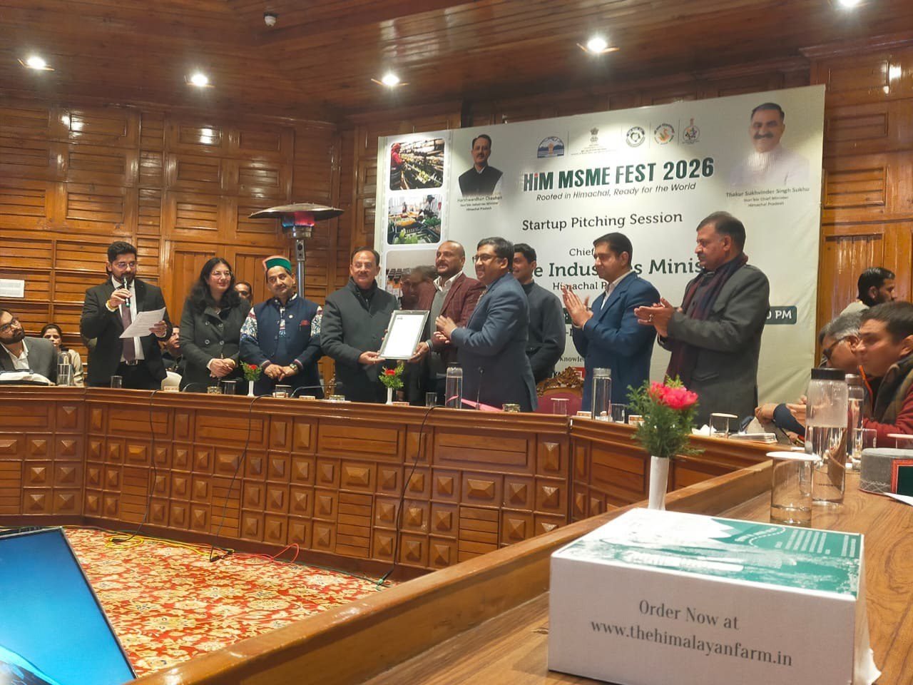 A framed certificate being presented at the HiM MSME Fest 2026 Startup Pitching Session. Two men hold the certificate between them in front of a long wood-paneled dais while officials and delegates applaud on either side. The event banner behind reads 'HiM MSME FEST 2026 — Rooted in Himachal, Ready for the World, Startup Pitching Session', with the Industries Minister named as chief guest.