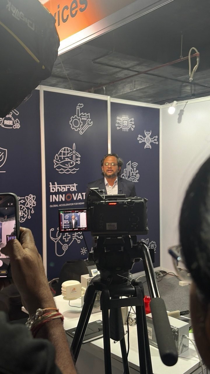 Professor K.V. Uday speaking to camera at Bharat Innovates 2026 at IIT Bombay. A broadcast camera on a tripod is trained on him in the foreground and another person films on a phone. Behind him is a dark blue backdrop printed with the Bharat Innovates logo and line-drawn icons of industry, computing, healthcare and energy, with hardware set out on the display counter.