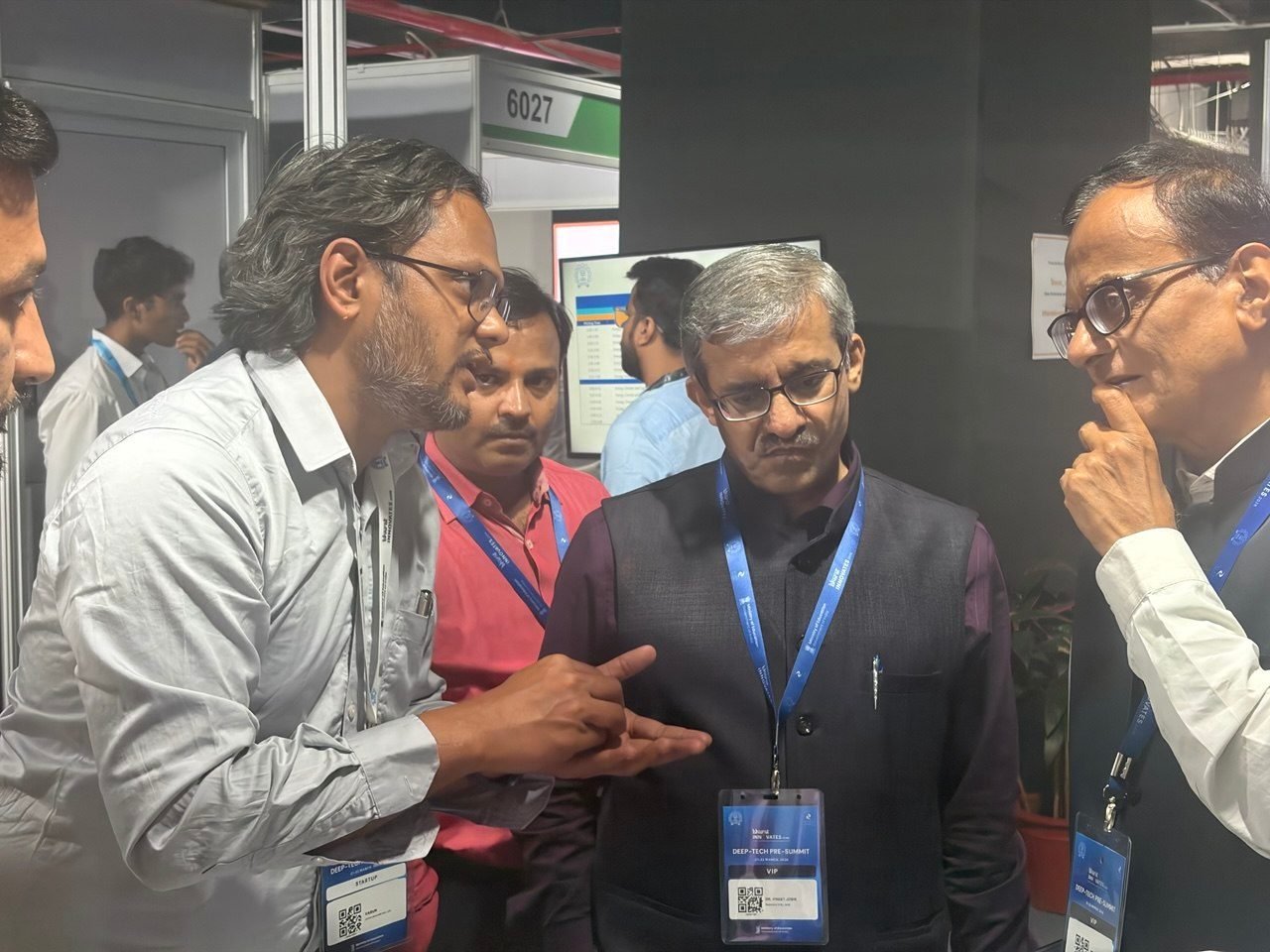 Professor K.V. Uday, in a white shirt with an exhibitor badge, leaning in and explaining something with his hands to two senior delegates wearing VIP lanyards at the Bharat Innovates Deep-Tech Pre-Summit. A colleague listens beside him, and a screen showing a table of readings is visible behind, next to stand number 6027.