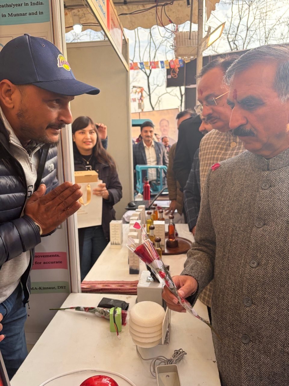 The Chief Minister of Himachal Pradesh, in a grey herringbone jacket, leaning in to look at iIoTs hardware on an exhibition counter while a member of the team greets him with folded hands and explains the system. A white stacked-disc sensor shield, a red warning beacon, a battery pack and cabling are set out on the table, with project posters behind the stand.