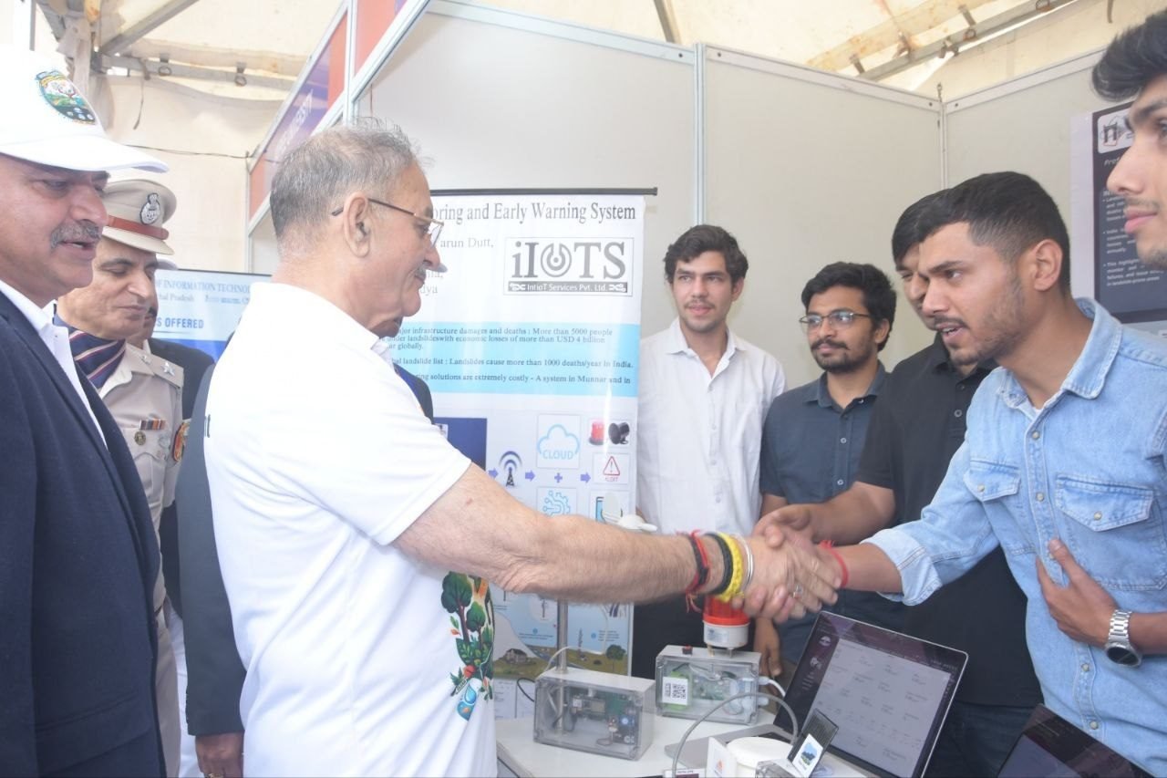 The Governor of Himachal Pradesh shaking hands with a member of the iIoTs IoT and server-side team at the company's World Environment Day stand in Shimla. Three more young engineers stand alongside; a uniformed police officer and another official are at the left. On the counter are an open sensor node in a clear enclosure, a red warning beacon and a laptop showing the monitoring dashboard, with the iIoTs Landslide Monitoring and Early Warning System banner behind.