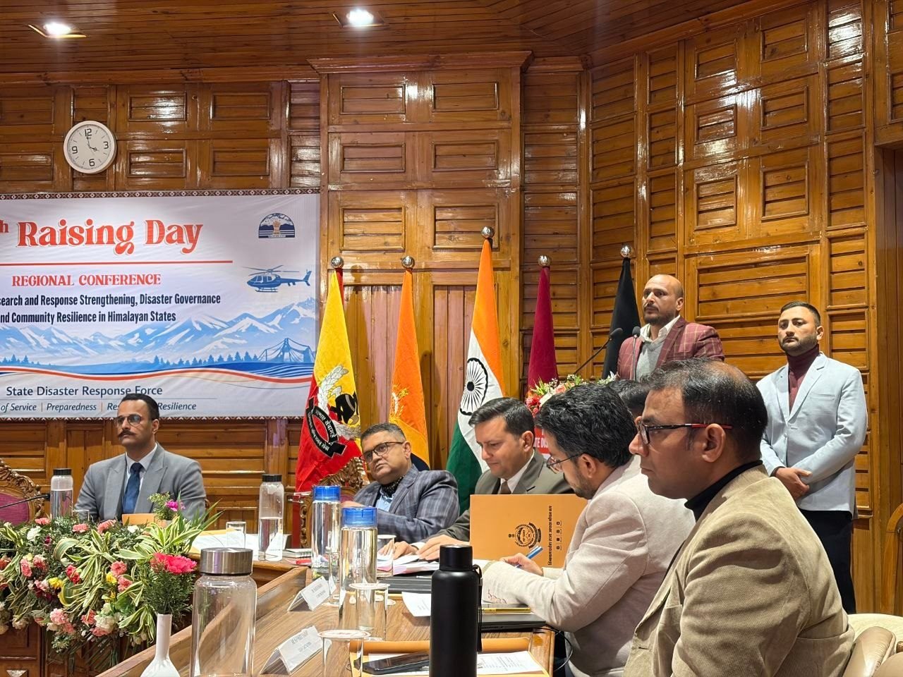 Chandan Vaidya of iIoTs speaking at a microphone in a wood-panelled conference room, addressing a seated row of officials in suits and uniform at the Himachal Pradesh State Disaster Response Force Raising Day regional conference, with the Indian tricolour and SDRF regimental flags behind them and the printed conference banner to the left.