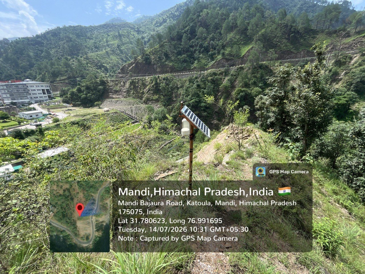 A compact solar-powered sensor unit on a short pole on a steep grassy slope above the Mandi–Bajaura road. A white instrument shield and an angled solar panel sit at the top of the pole. Terraced fields, the road, and the buildings of IIT Mandi's north campus are visible across the valley, with forested ridges beyond. A GPS camera stamp in the corner records the coordinates and the date.