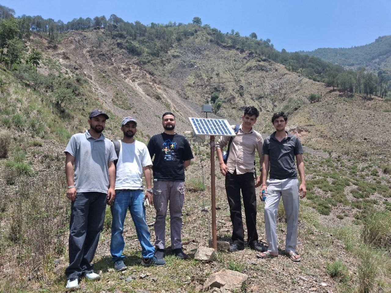 Five members of the iIoTs team standing beside a newly installed monitoring station at Kotropi — a steel pole carrying a solar panel, an instrument shield and a sensor enclosure, set in rock at their feet. Behind them the hillside is stripped bare across its whole width by a landslide scar, with surviving pines along the ridge above it.